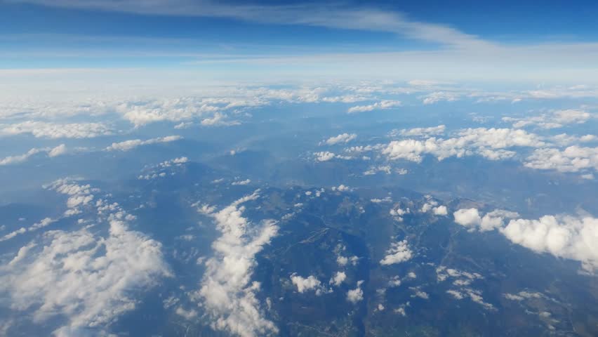 Top view of mountainous terrain with partial cloudiness above her and transversal contrail by another plane, view from an airplane side window during a flight at high altitude
