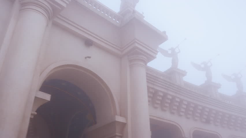 Columns and angel Statues rising through thick Fog in a Wide Angle architectural view at Ba Na Hills showing soft daylight and pale atmospheric tones.