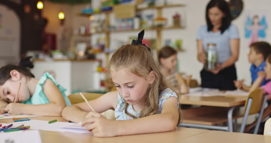 Focused elementary school girl writing at desk in classroom, doing homework or test, studying with classmates while teacher helps other children in the background.