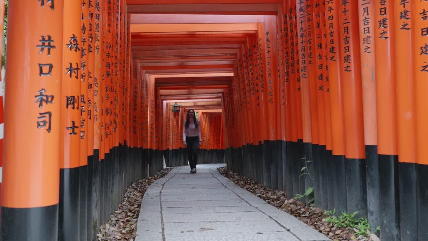 Young caucasian woman walking through a stone path surrounded by red and black wooden torii gates at Fushimi Inrai Taisha, Kyoto, Japan.