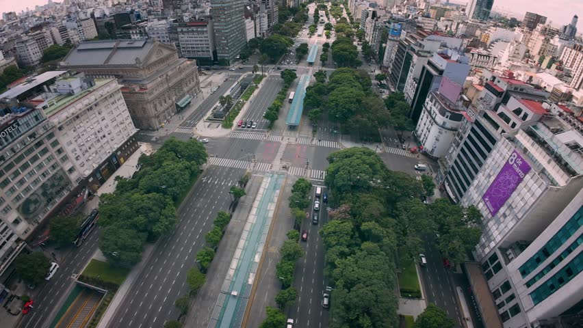 Aerial drone view of Avenida 9 de Julio in Buenos Aires, Argentina, featuring the iconic Teatro Colón, wide multi-lane avenues, tree-lined medians, modern city traffic, and dense urban architecture. This cinematic cityscape showcases one of the widest avenues in the world and reflects the scale, culture, and metropolitan life of Argentina’s capital city.
