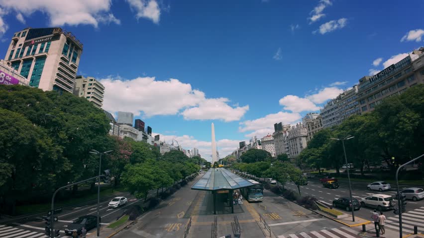 Aerial drone view of the Obelisk of Buenos Aires rising above Avenida 9 de Julio, one of the widest and most iconic avenues in the world. This cinematic shot captures the symmetrical layout of the avenue, the dedicated bus lanes of the Metrobus system, dense urban architecture, and lush green trees framing the city’s central axis. The Obelisk stands as a historic and cultural landmark, symbolizing the identity, scale, and rhythm of Buenos Aires, Argentina.
The footage showcases modern urban infrastructure combined with historic buildings, active traffic flow, and organized public transportation corridors. Shot in daylight under a clear blue sky with scattered clouds, this aerial perspective highlights the contrast between nature and architecture in a major South American capital. The smooth drone movement emphasizes depth, scale, and urban geometry, making it ideal for cinematic storytelling.
This clip is well suited for commercial, editorial, tourism, and documentary projects related to travel, city branding, transportation systems, smart cities, urban development, and Latin American destinations. It represents contemporary city life, mobility, and architecture in Buenos Aires, one of the most important metropolitan centers in the region.
