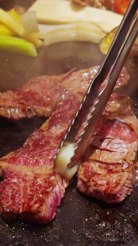Vertical video. Searing Beef Strips Browning on Hot Grill Beside Vegetables as Tongs Lift Meat Through Rising Steam.