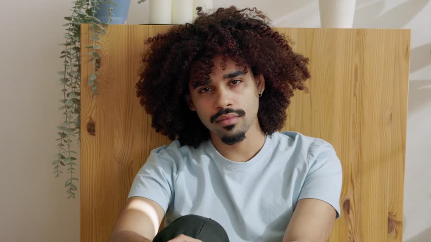 Portrait of young handsome Latin American man with a stylish afro haircut sitting at home in the morning sunshine, looking at the camera and showing confidence and positive attitude