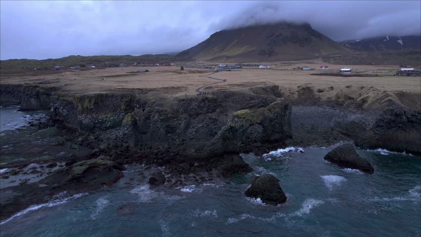 Aerial view of a remote coastal village in Iceland with volcanic cliffs, rugged shoreline, and ocean waves. Mountain partially covered by low clouds creates a moody Nordic landscape.