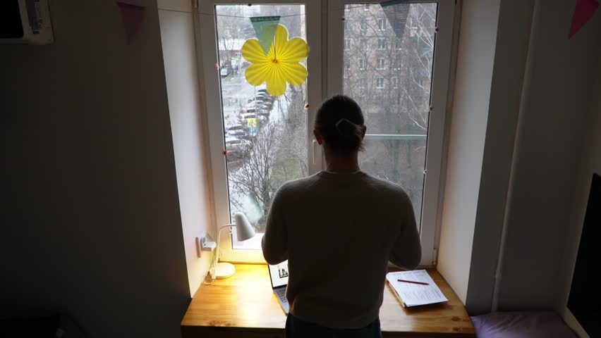 High angle back view of a person leaning over a wooden desk and looking out of a window in a home office, with soft natural light illuminating the scene and papers scattered on the workspace