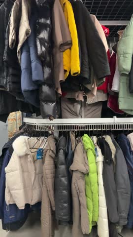 Rack of winter coats in store packed with colorful puffer jackets and wool coats on metal hangers, two-tier rail under grid ceiling, closeup of textured fabrics and zippers,