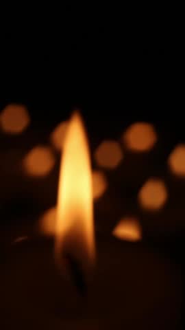 A candle flame appears in close view as it moves slightly upward across the frames while distant warm bokeh lights behind it shift gently, creating subtle background motion