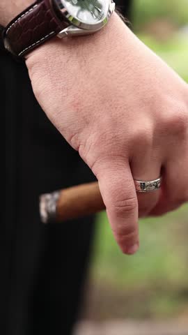 Close-up of groom or man hand in pinstripe suit holding lit cigar, wristwatch visible. Elegant outdoor moment of relaxation and celebration. A man is holding a cigar.