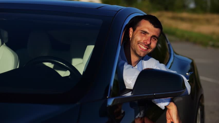 Young smiling entrepreneur leaning out of driver window at modern electric car and looking into camera. Portrait of handsome successful businessman showing thumbs up after bought new electrical auto