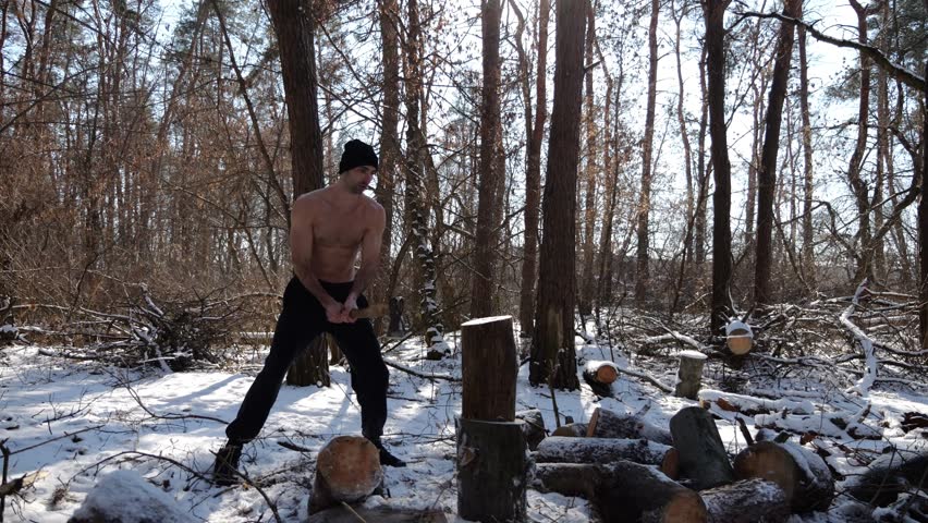 Shirtless young man chopping firewood in a sunny winter forest. Muscular guy working with axe in snowy woodland. Concept of strength, masculine, energy and harmony with nature during cold season