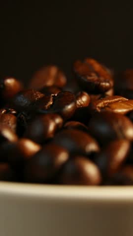 Dark roasted coffee beans subtly shift position as focus transitions across the pile under warm soft studio lighting creating slow tactile motion highlighting texture and glossy surfaces