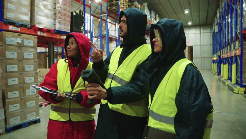 Three diverse colleagues in warm uniforms checking inventory with a barcode scanner inside a large, refrigerated industrial warehouse