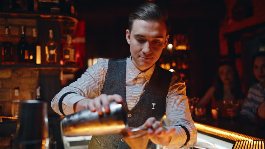 Focused bartender pouring ingredients from shaker to elegant glasses at bar counter closeup. Skilled professional barman preparing alcoholic cocktails to club customers. Young club worker making drink