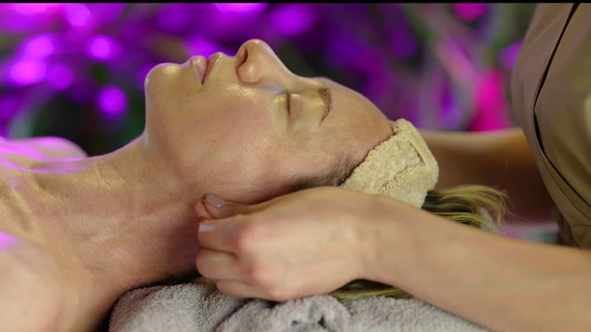Relaxed middle aged woman receiving treatment in beauty salon, professional lady masseur making massage for head and face, gently massaging ears for female client, wellness and body care routine