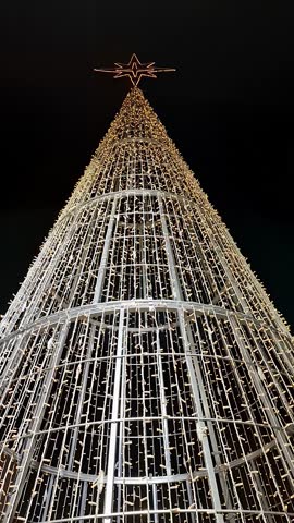 An illuminated christmas tree stands tall during the night, showcasing a unique avant garde design. Bright lights cover the tree, creating a striking visual display for all to see.