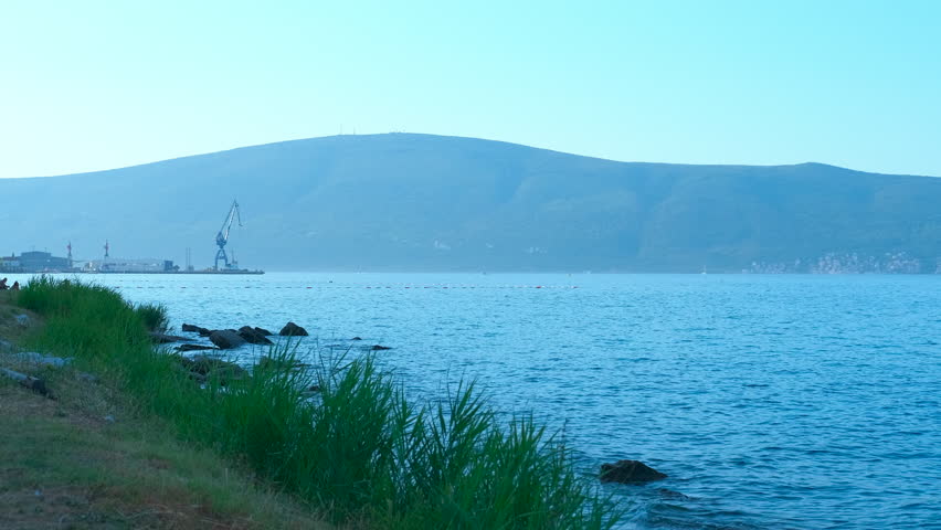 Peaceful coastal landscape with sea and mountains. Calm sea waves gently lapping against a rocky shore, with mountains and a distant port visible in the hazy background on a clear day