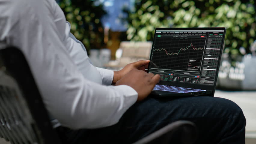 Black man navigating stock trading indicators online on laptop, person seated on a bench works outdoors in a park. Metropolitan area with skyscrapers and sidewalk motion, urbanism. Camera B.