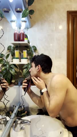 Young man takes care of his appearance, shaving his beard with an electric razor in front of a bathroom mirror