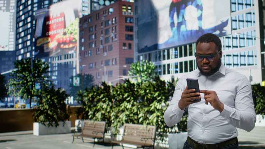 Black man looking for directions on smartphone map app during a stroll, walking through urban street with office buildings. Vibrant city life within a metropolitan city center area. Camera A.