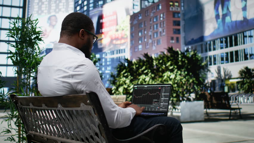 Dedicated adult man monitoring stock market exchange trends on laptop, working remotely on laptop from a bench outdoor. Sitting in a small park downtown and analyzes funds. Camera A.