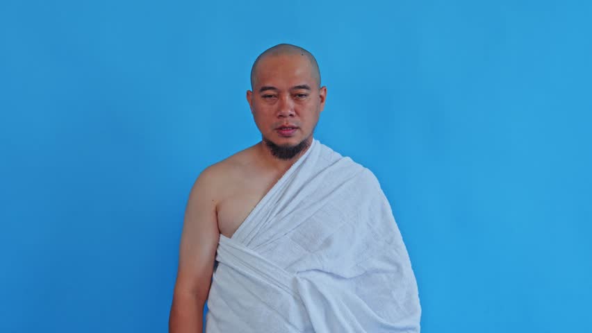 Devout Muslim man in Ihram performing sincere pray with closing face-wipe gesture isolated on blue background