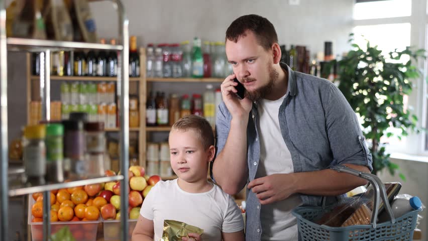Dad and son, while shopping for groceries, call up with mom, discuss on phone purchase of snacks for festive lunch. Husband informs his wife on phone about cost of groceries in supermarket