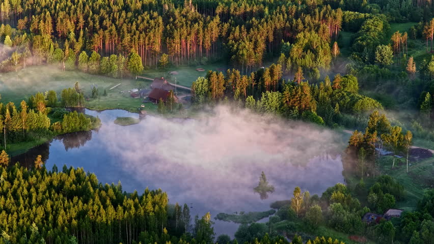 A serene morning aerial view of a forest lake with low mist drifting across the water, surrounded by dense green trees and a small wooden cabin near the shore.