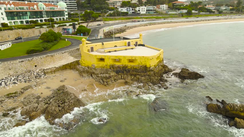 Aerial drone footage of Forte da Giribita and rocky coastline in Caxias Portugal with ocean waves hitting stone reefs beside a coastal highway and seaside promenade on a cloudy day.
