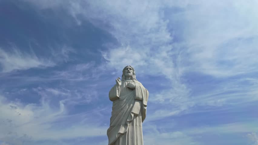 Tilt-down shot of the Áldó Krisztus szobor statue in Tarcal, Hungary, surrounded by serene hills, greenery, and clouds, showcasing its religious and cultural significance in a scenic landscape.