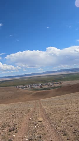 Vertical video of a car driving along a country road under blue sky. Altai desert and semi-desert environment. The village of Kokorya is visible in the valley.