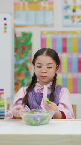 Smiling Asian schoolgirl eats fresh vegetable salad at classroom desk with bright colorful background