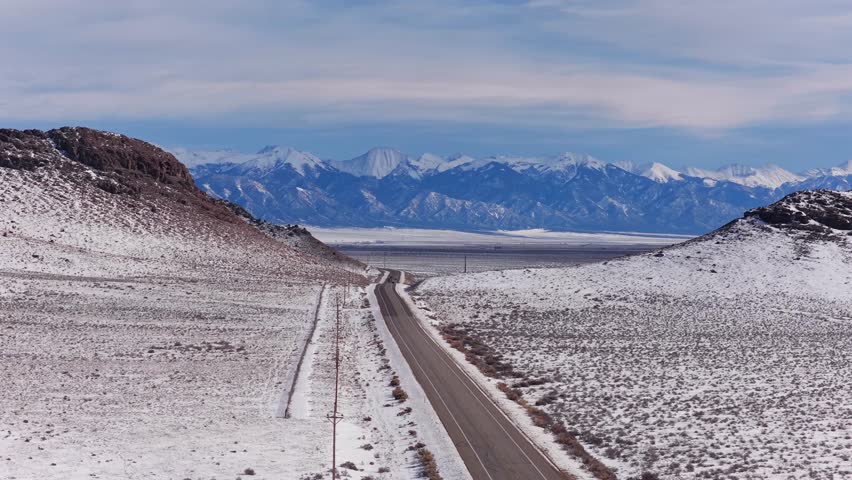 Aerial perspective of a remote highway and wide, snow-dusted arid plain, defined by sparse vegetation and flanking hills. The road leads to the massive Sangre de Cristo range in Colorado.