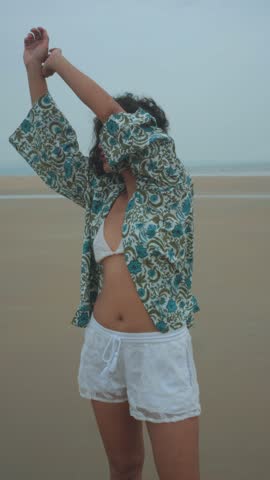 A young woman stretching on the beach, wearing a patterned shirt, white bikini top and lightweight shorts, captured in a relaxed and breezy summer atmosphere.