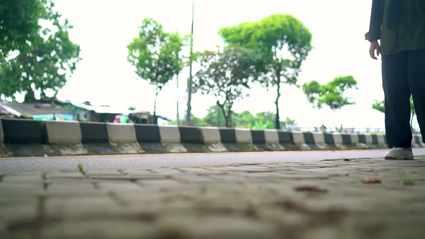 Low angle view of a person walking along a city sidewalk beside a road, showing casual movement, footwear steps, and urban surroundings with trees and passing vehicles in daylight.