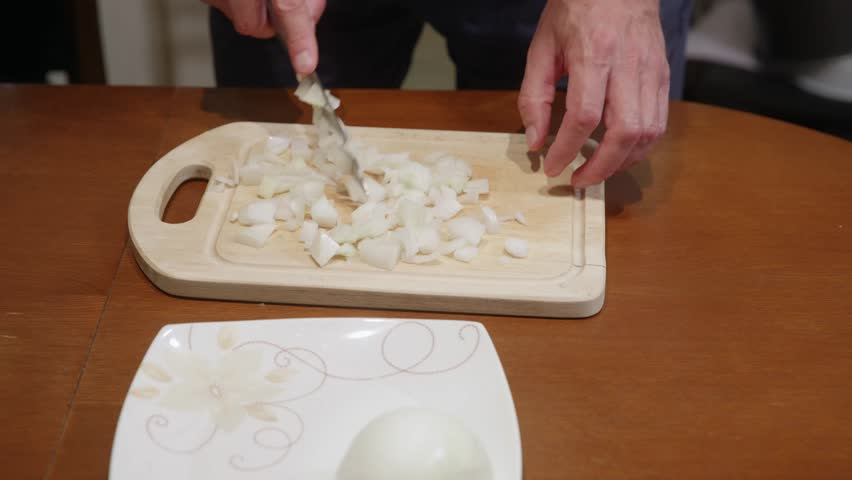 Person putting peeled onion bulb on natural wood board with decorative white plate nearby on brown wooden table. Food preparation scene showing fresh vegetable ingredient for cooking.