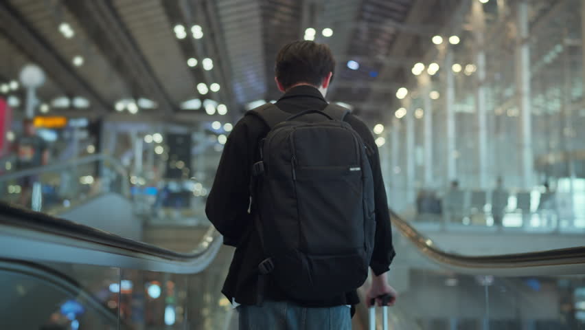 Rear view of young male traveler with black backpack and suitcase walking on escalator at international airport terminal for travel and vacation concept