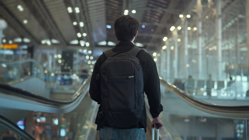 Rear view of young male traveler with black backpack and suitcase walking on escalator at international airport terminal for travel and vacation concept