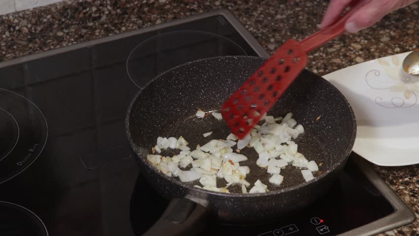 Diced white onion sauteing and browning in dark speckled non-stick pan on induction cooktop. Cooking process showing caramelization of vegetable ingredient for homemade meal preparation.