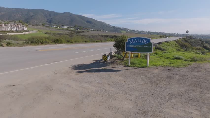 An aerial shot captures the Malibu CA sign as the camera moves smoothly from left to right, revealing the surrounding landscape and Pacific coastline under a clear sky.
