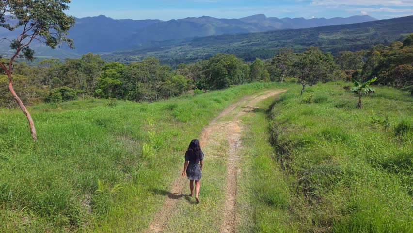 A drone follows a woman walking along a grassy path, framed by green trees and mountain landscapes under bright sunlight. A calm, cinematic scene evoking freedom, travel, and nature.