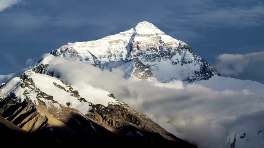 Mount Everest stands tall amidst a cloak of clouds, highlighting the dramatic interplay of light and shadow as the sun rises, capturing the breathtaking beauty of nature.