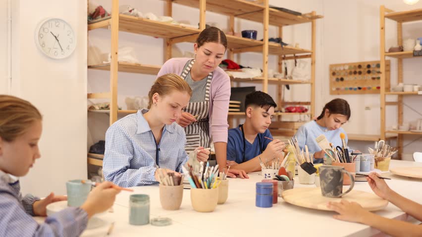 Children and teenagers sit at a table and paint ceramics at the workshop. Teacher shows how to paint a clay product after firing
