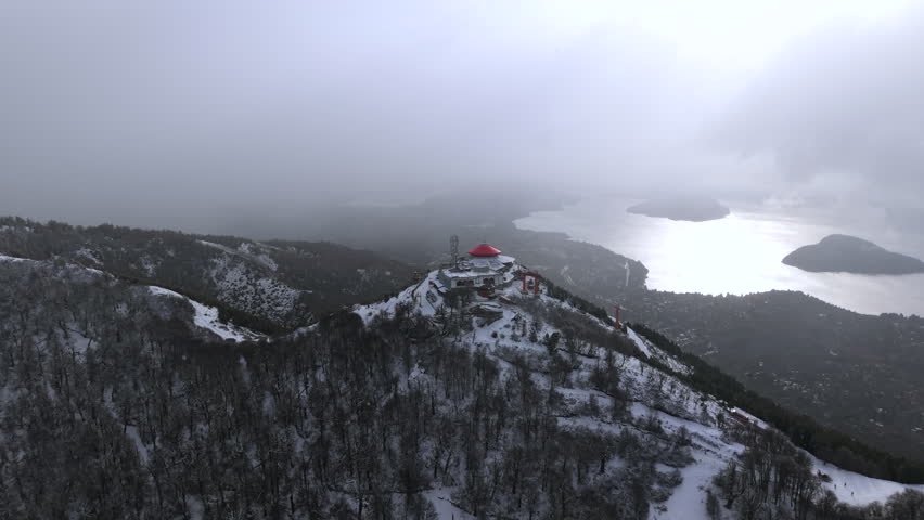 Snowy Cerro Otto ridgeline leading to red-roof summit building above Bariloche lakes under stormy sky.