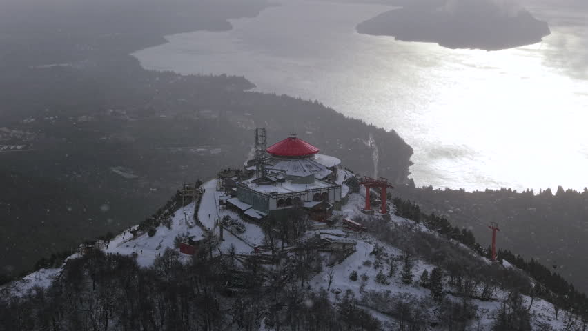 Snowy Cerro Otto summit with red-roof building and towers overlooking Bariloche lake during winter storm.