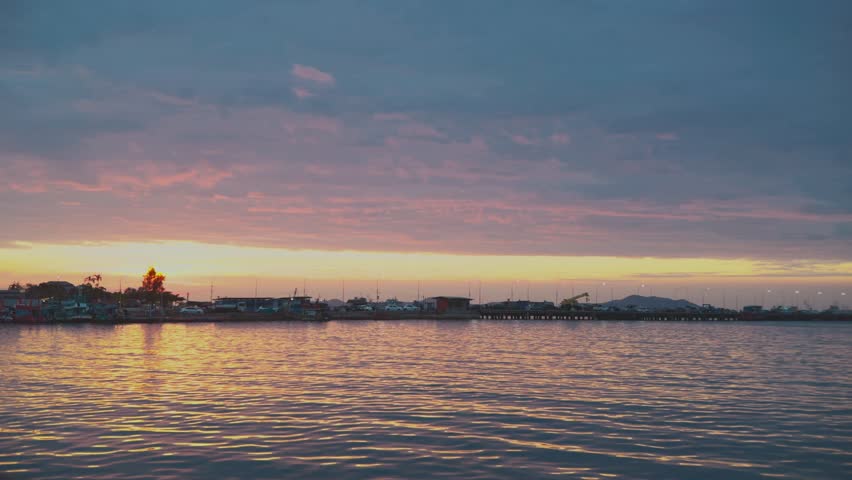 sunset over calm ocean water, pink and gold sky reflecting on rippling surface, low distant coastline and small pier silhouette,