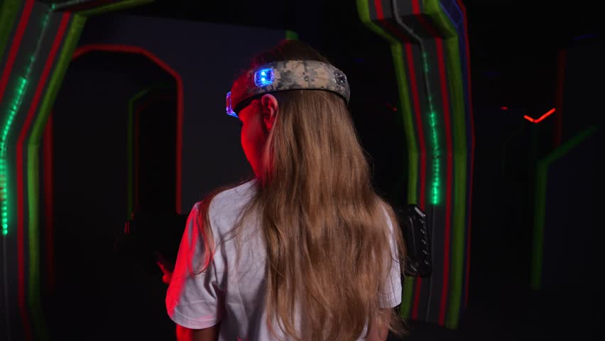 Portrait of an active girl enthusiastically playing laser tag in a dark room with neon stripes on the wall. A girl with a laser gun and a marker on her head is playing laser tag.