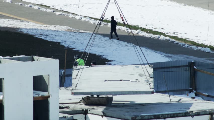 Overhead view of construction site action with two workers engaged in guiding a heavy, rectangular precast concrete slab. Crane machinery lowers the massive building material
