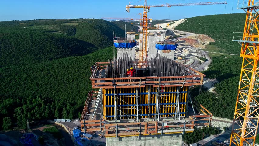 Drone view of workers assembling steel reinforcement and formwork on a reinforced concrete bridge pier using tower crane at a major highway construction site. Large-scale infrastructure and civil engineering project.