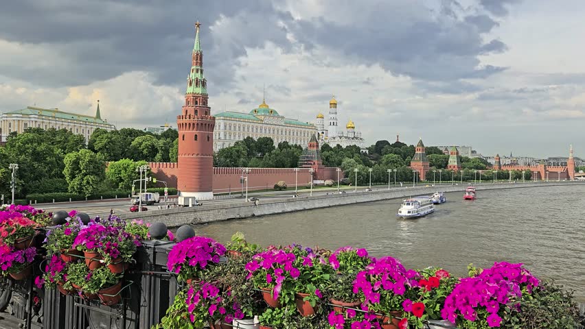 Boat is traveling down a river near a city Moscow. The river is lined with Kremlin wall, buildings and trees. The bridge has a row of potted plants with flowers